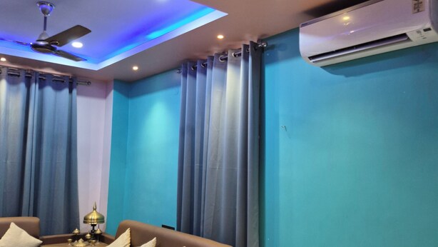 A hotel room with a bed, a sofa, a nightstand, an air conditioner, a ceiling fan, and curtains. - Cozy Roost, quiet place (Guwahati)