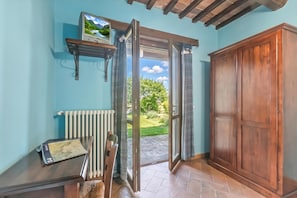 A room with a wooden door, a radiator, a shelf with a picture frame, and a view of greenery outside.