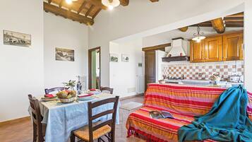 Un coin repas avec une table dressée pour deux personnes, un canapé avec une couverture, des poutres en bois au plafond et une cuisine en arrière-plan.