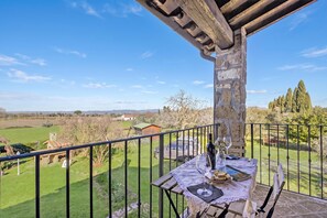 Un balcone con un tavolo apparecchiato per consumare un pasto, con vista su un paesaggio rurale ricco di campi e alberi.