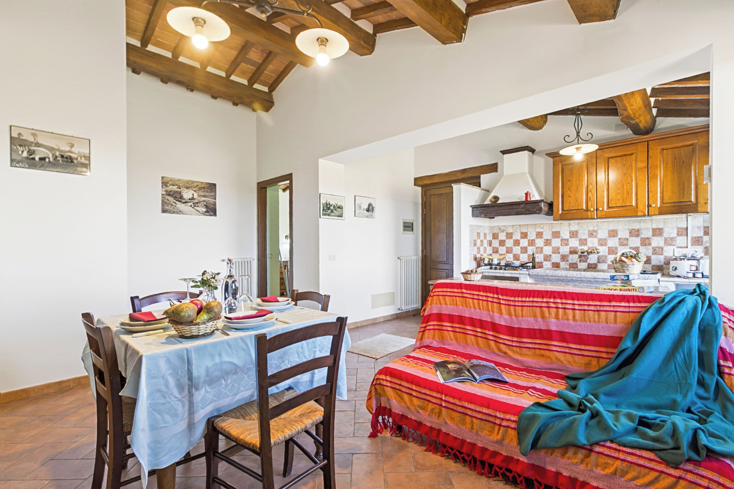 Un'area pranzo con un tavolo apparecchiato per due, un divano con una coperta, travi in legno al soffitto e una cucina sullo sfondo.