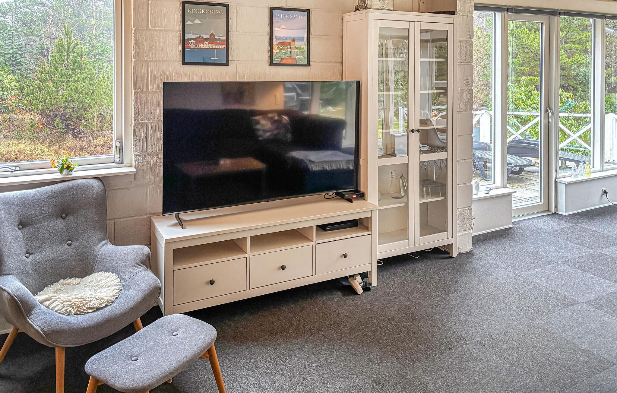 Olohuoneessa on litteänäyttöinen televisio, harmaa nojatuoli, pieni jakkara, lasiovilla varustettu kaappi ja ikkuna, josta avautuu näkymä vehreään maisemaan.