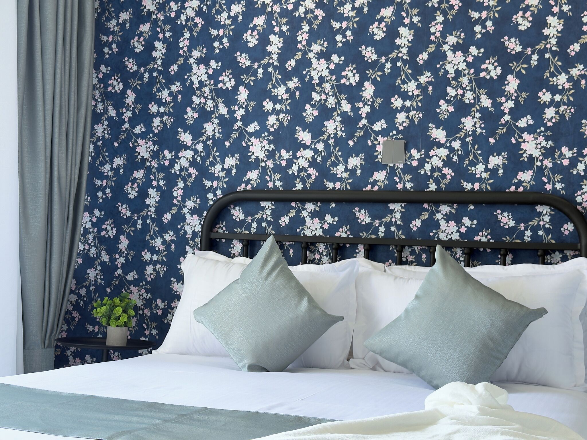Un lit avec un cadre en métal, une literie blanche et deux oreillers bleu clair, le tout sur un fond de papier peint à motifs floraux.