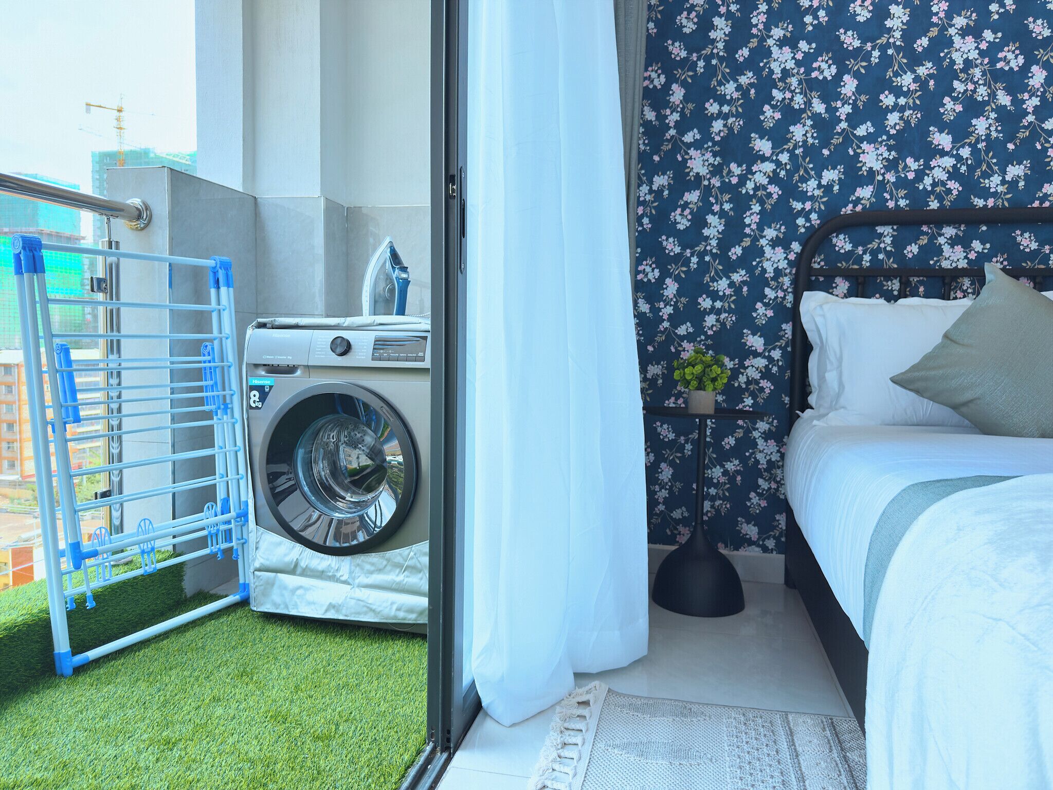 A room with a bed, a washing machine, a drying rack, and a small potted plant.