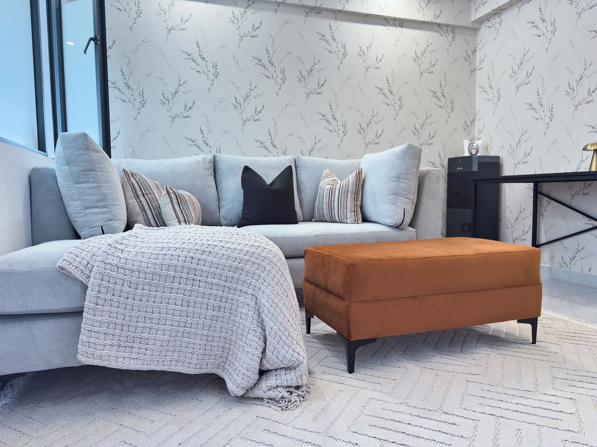 A modern living room with a grey sofa, a patterned throw blanket, a brown ottoman, and a herringbone-patterned rug.