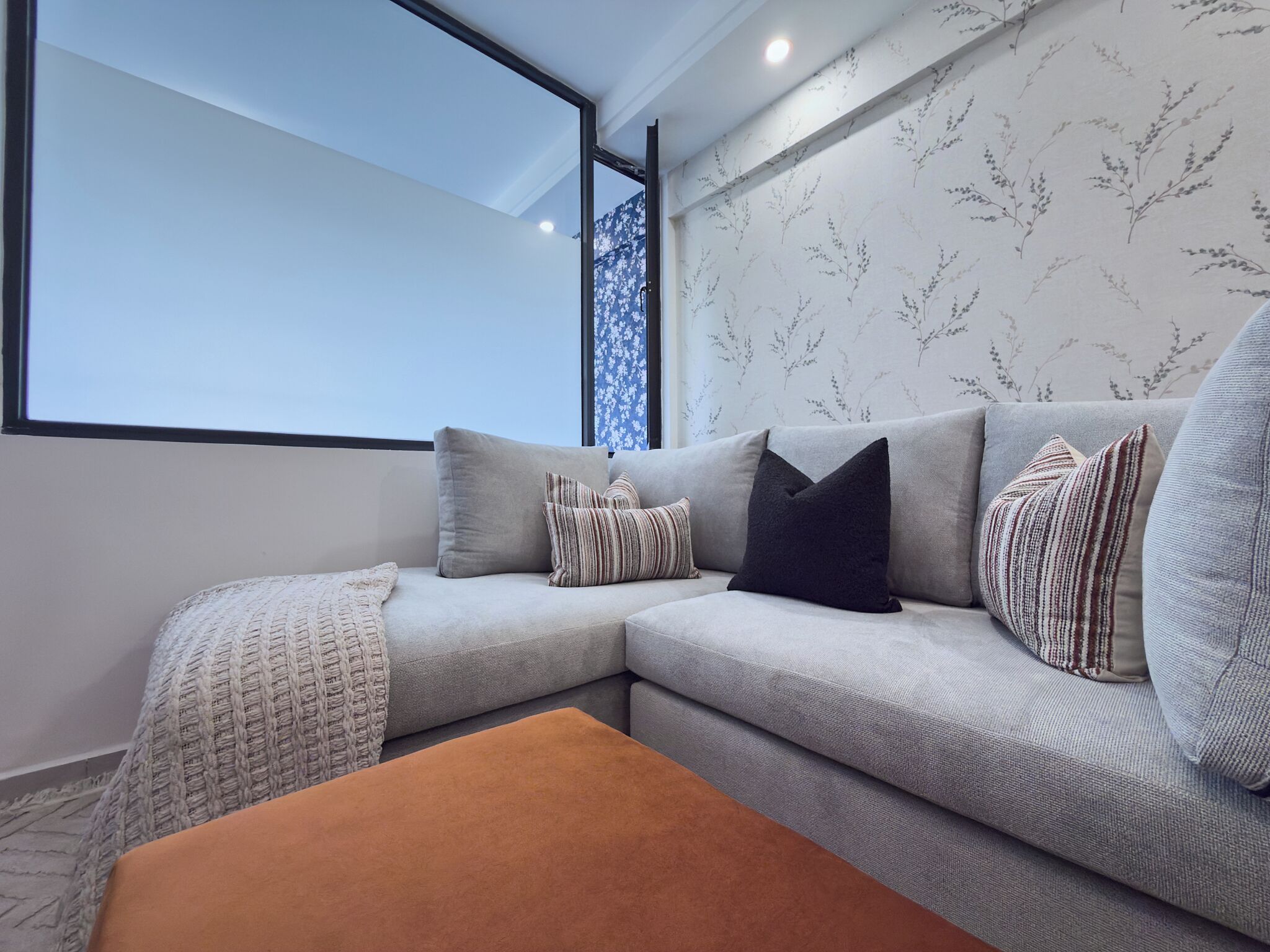 Un salon moderne avec un canapé d’angle gris, des coussins à motifs, un plaid tricoté et un pouf marron.