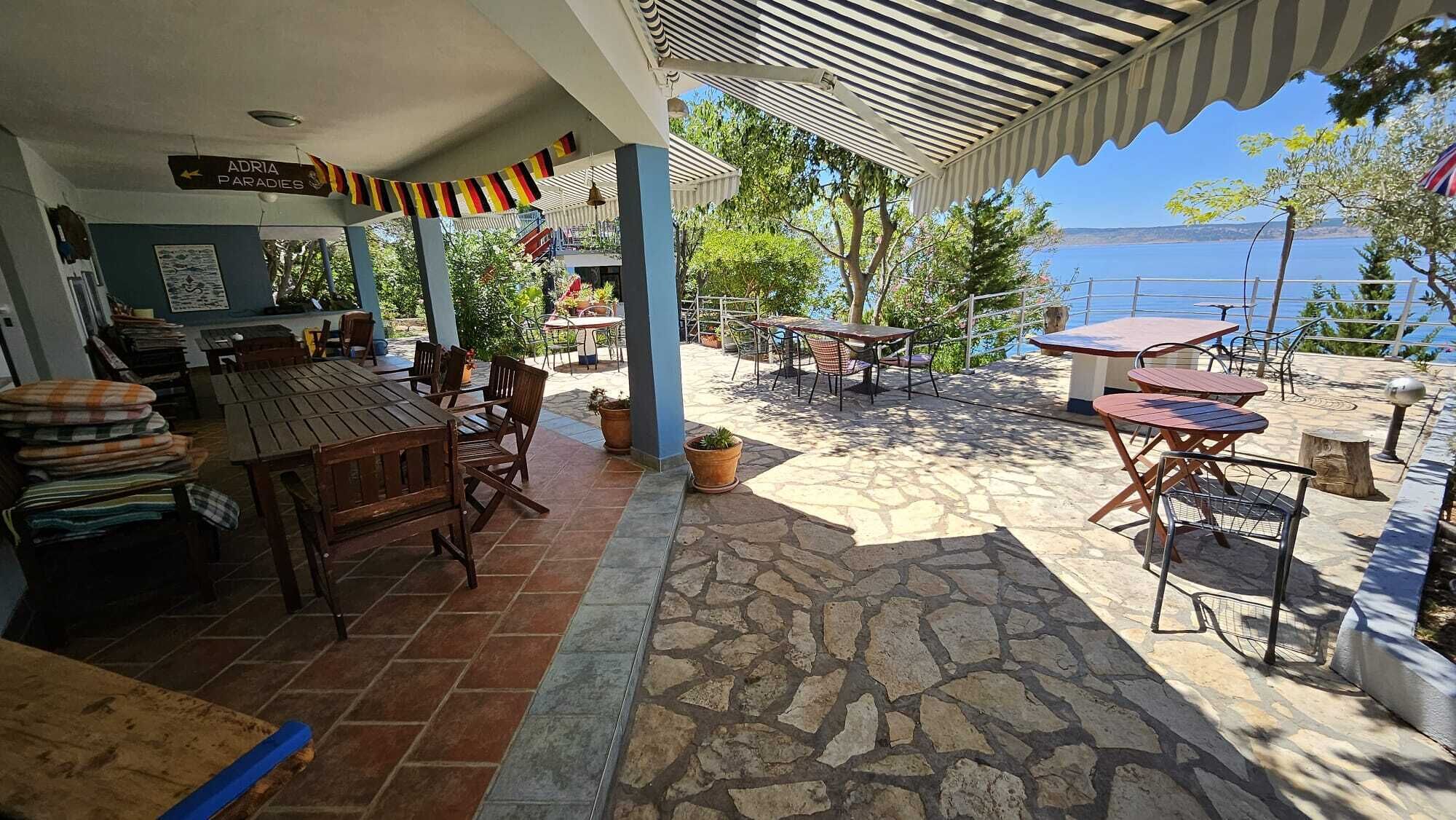 Un'area salotto all'aperto riparata, con pavimento in pietra, tavoli e sedie in legno e vista sul mare.