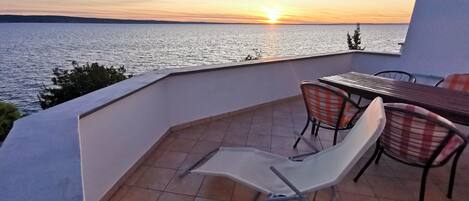A balcony with a view of the sea at sunset, a table, and two chairs.