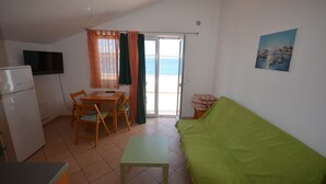 Ein kompakter Wohnraum mit einem grünen Sofa, einem Esstisch mit Stühlen, einer kleinen Küchenzeile inklusive Kühlschrank und einer Glastür zum Balkon.