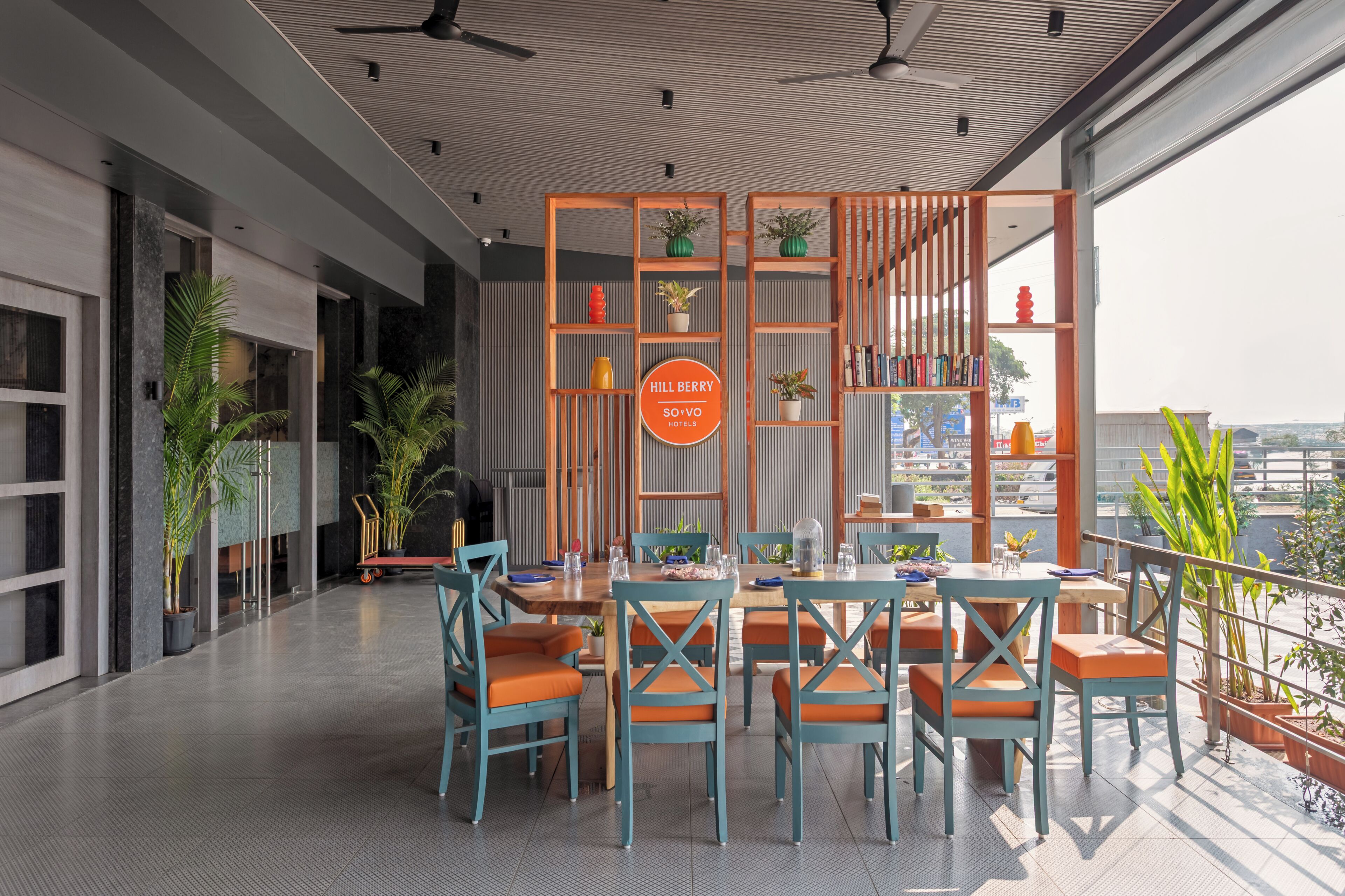 A modern hotel lobby with a dining area, potted plants, and a sign for Hill Berry So'Vo Hotels.