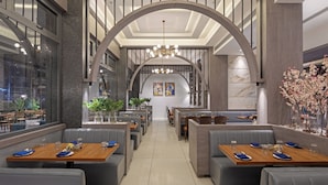 A modern restaurant with high ceilings, arched windows, and a central chandelier. Tables are set with blue napkins and plates. - Hill Berry - A SoVo Hotel (Mawal)