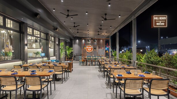 A restaurant with multiple tables and chairs, ceiling fans, and a view of the city at night. - Hill Berry - A SoVo Hotel (Mawal)