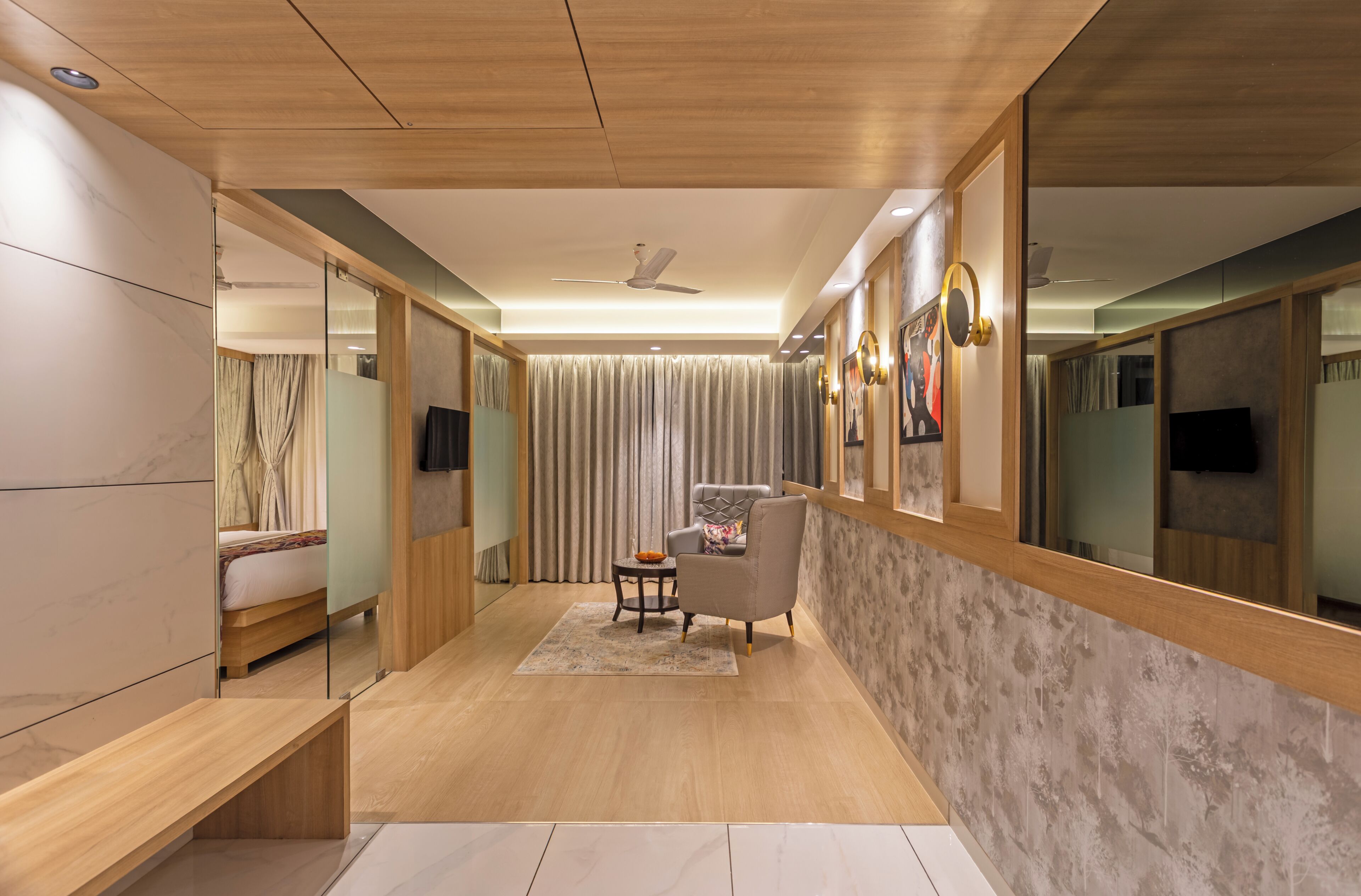 A modern hotel room with a wooden ceiling, a large mirror, a bed, a seating area with a chair and table, and a television.
