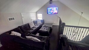 A living room with a black leather sofa, a small table, and a television mounted on the wall. - NONA Luxe 5BR 4BH  w/ Guesthouse / Theatre / GameRoom / Gym / Loft (Unknown)