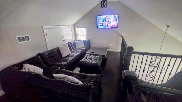 Uma sala de estar com sofá de couro preto, uma mesa pequena e uma televisão fixada na parede.