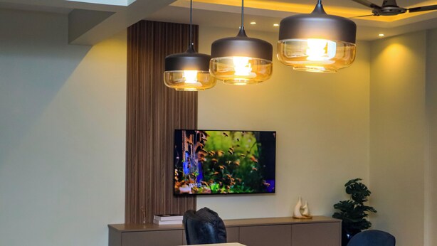 A modern dining area with a long table, two pendant lights, a ceiling fan, and a flat-screen TV mounted on the wall. - Norden Suites (Mombasa)