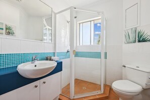 A bathroom with a white sink, blue countertop, white toilet, and a glass-enclosed shower. - Oceanside Apartment with Views of Yaroomba Beach (Point Arkwright)