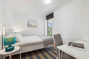 Ein kleines, sauberes Zimmer mit einem Bett, einem Schreibtisch, einem Stuhl, einer Lampe, einem Fenster und einem Bild an der Wand.