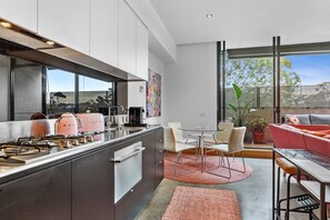 Eine moderne Küche mit Edelstahlgeräten, ein Essbereich mit Glastisch und Stühlen und eine gemütliche Sitzecke mit einem rosafarbenen Sofa und einer Pflanze.