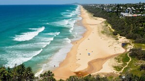 Una playa costera con orilla arenosa, agua azul cristalina y un área residencial al fondo.