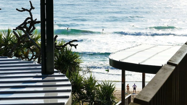 Vista de la playa desde un área techada con barandilla, plantas de palma y gente practicando surf en el océano.