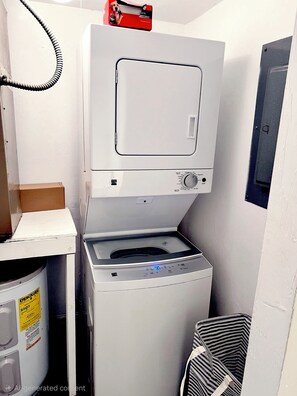 A compact laundry area with a stacked washer and dryer, a small cabinet, and a trash bin. - Charming Budget-friendly 1BR Stay in Gainesville (GAINESVILLE)