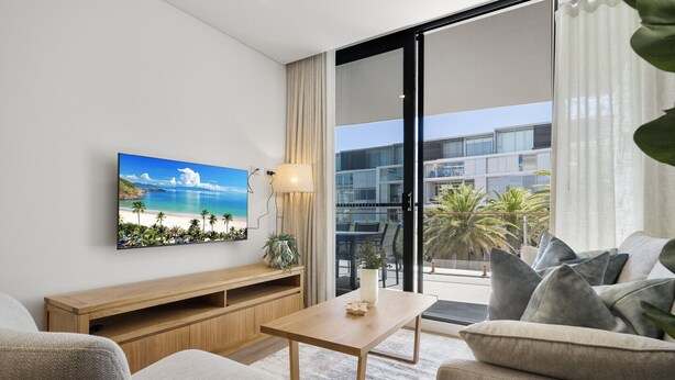 A modern living room with a flat-screen TV, wooden furniture, a balcony with a view of a building and palm trees. - Fremantle 1-Bed Close to Beach & Shops (North Fremantle)
