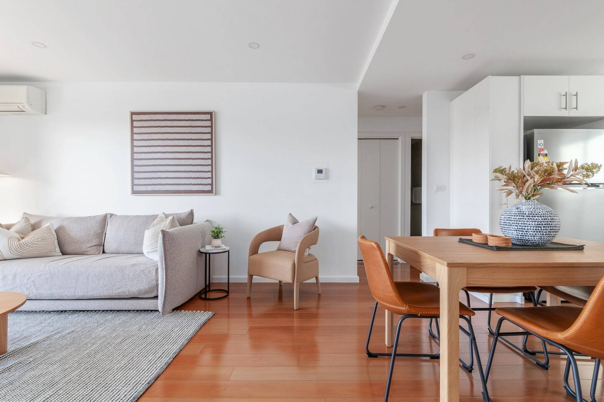 A modern living room with a sofa, a small table, a chair, a dining table with chairs, and a kitchen area with cabinets.
