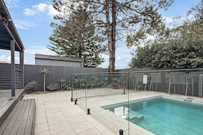 Une piscine avec une terrasse en bois, une clôture en métal et, au fond, un bâtiment au design moderne.
