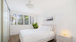 Un dormitorio con cama, mesita de noche, lámpara, ventana con persianas y una planta.