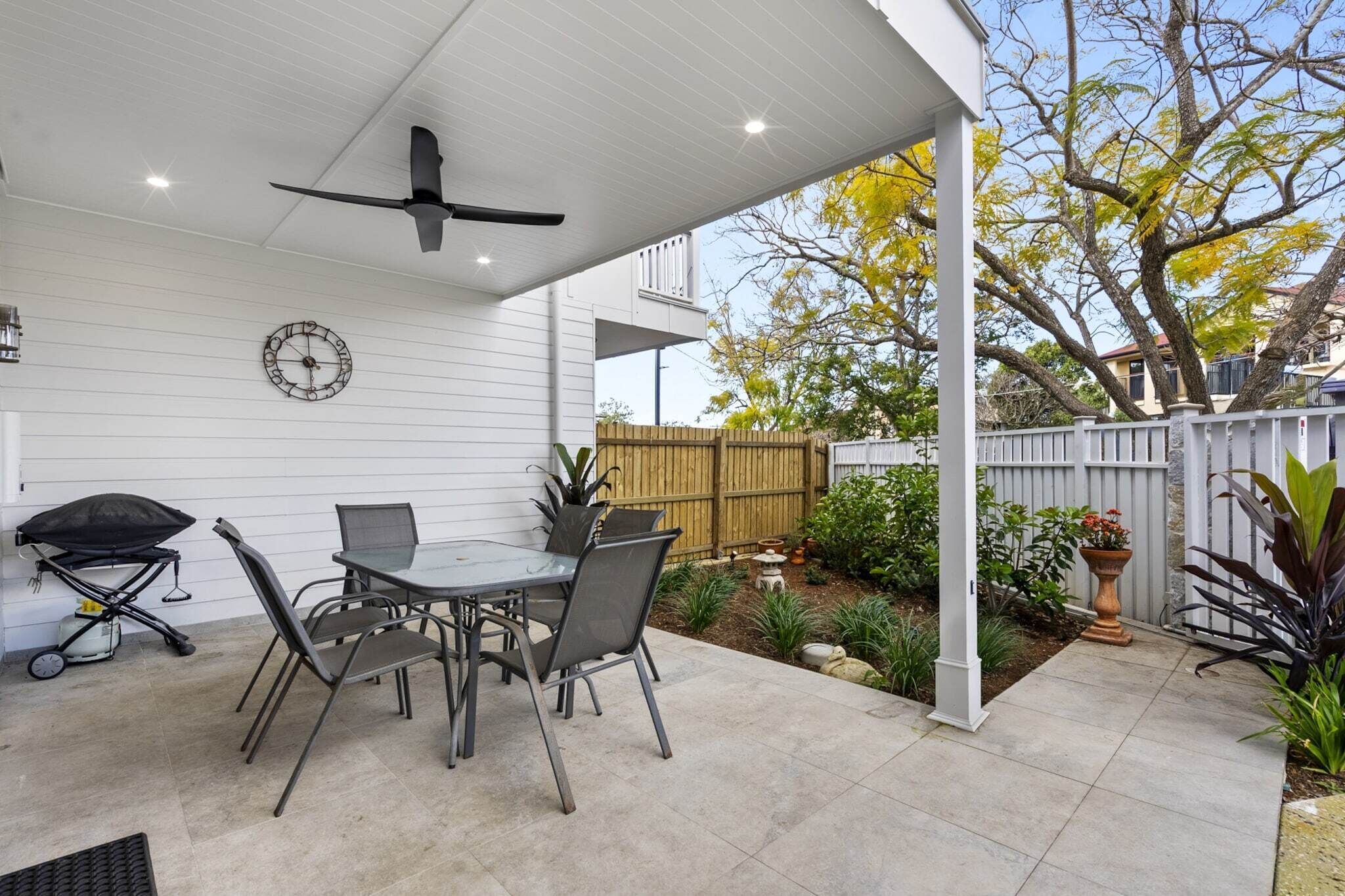A covered patio with a table and chairs, a ceiling fan, a clock, and a garden with a wooden fence.