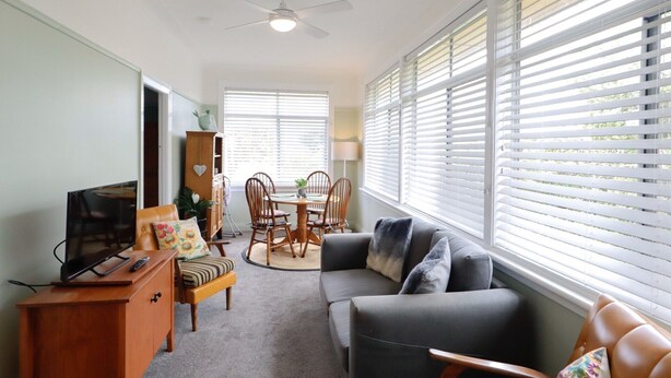 A living room with a TV, sofa, dining table, chairs, and large windows with blinds.