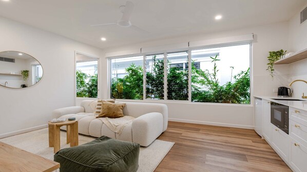 A modern living room with a sofa, a small table, and a view of greenery through large windows. - BAHA Breeze | Luxury Villa in Port Douglas (PORT DOUGLAS)