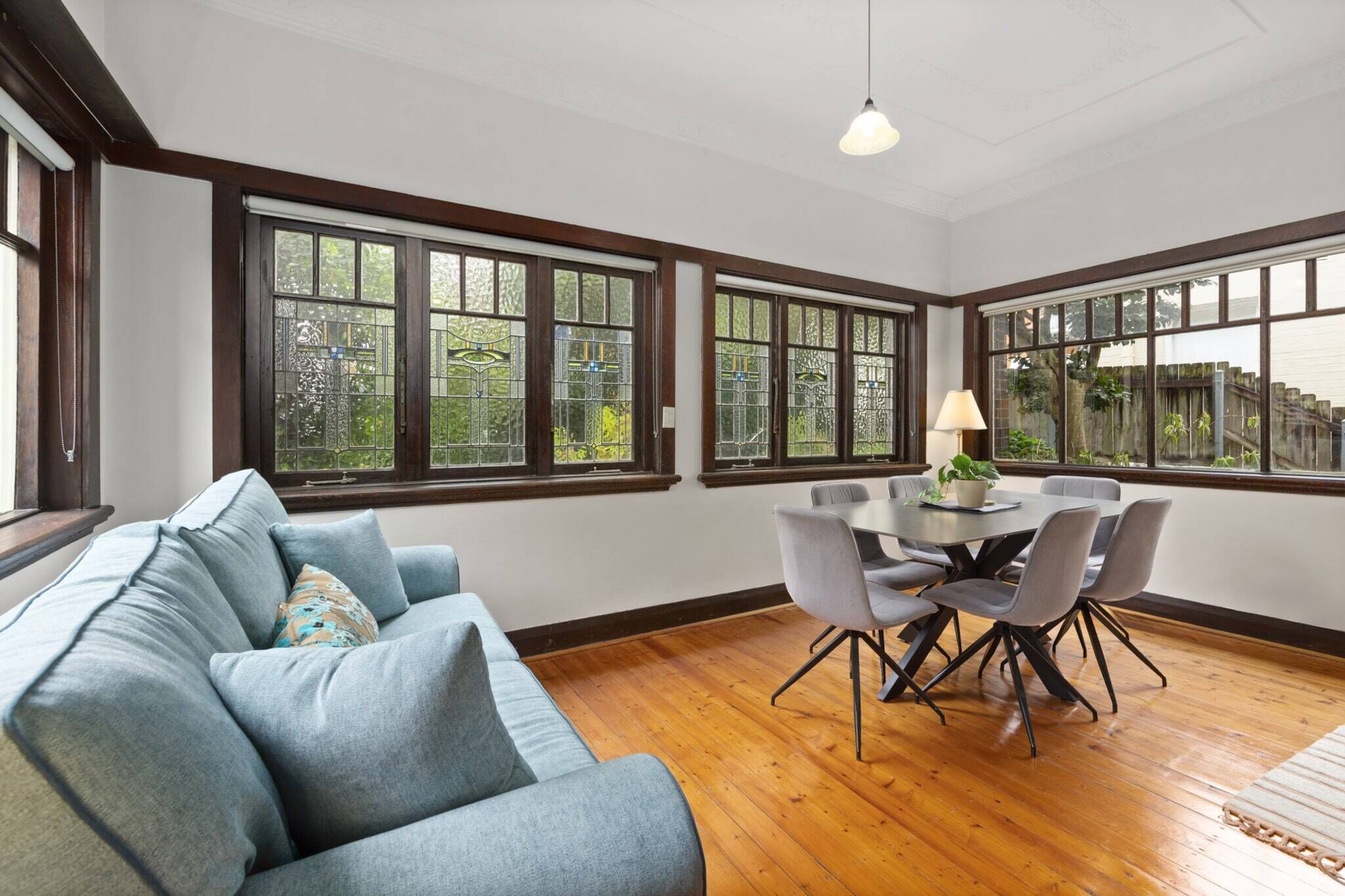 A room with a blue sofa, a dining table with chairs, and large windows with a view of greenery.