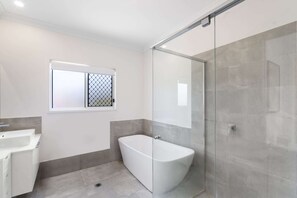 Un baño moderno con ducha envuelta en vidrio, una bañera independiente y un ventanal con celosía metálica.