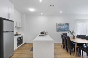 A modern kitchen with a central island, a dining area with a wooden table and chairs, and a painting on the wall. - Family-Friendly House with Courtyard Retreat (Wavell Heights)