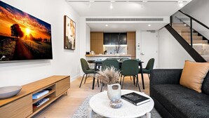 A modern living room with a flat-screen TV, a dining area with chairs, a kitchen with cabinets, and a staircase. - Stylish 3-Bed Townhouse in Vibrant Fitzroy (Fitzroy)