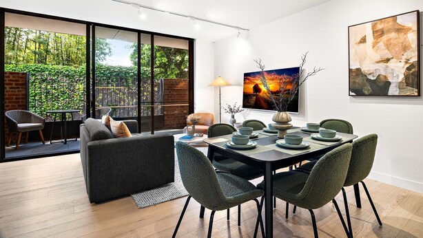 A modern dining area with a table set for four, surrounded by green chairs, a sofa, and a view of a brick wall and greenery outside. - Stylish 3-Bed Townhouse in Vibrant Fitzroy (Fitzroy)