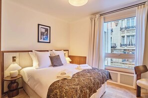 Ein Hotelzimmer mit einem großen Bett, einem Nachttisch mit Lampe, einem Fenster mit Blick auf Gebäude und einem gerahmten Bild an der Wand.