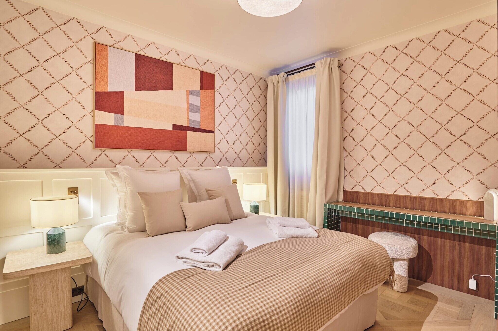 Habitació d'hotel amb un llit, una taula de nit amb una làmpada, un bany amb mirall i una finestra amb cortines.