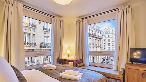 A hotel room with a large bed, a desk, a TV, and a view of Parisian buildings. - Sublim AC Apartment 3 Bdr Saint Germain des Prés (Paris)