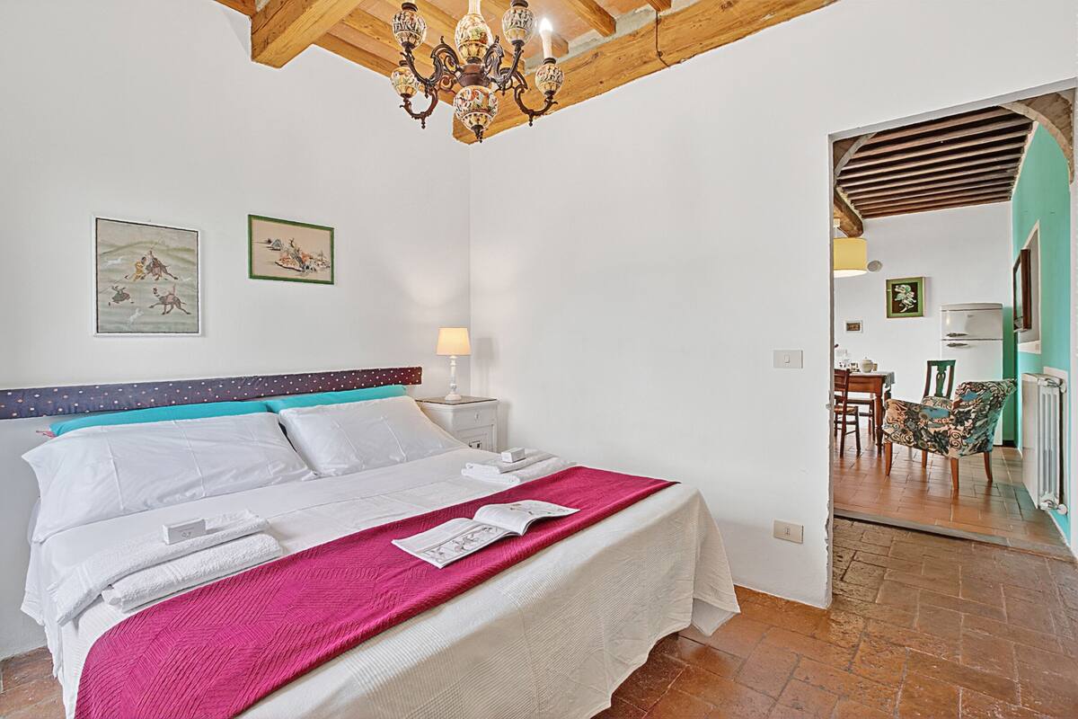 A bedroom with a bed, a chandelier, a nightstand, and a door leading to a dining area.