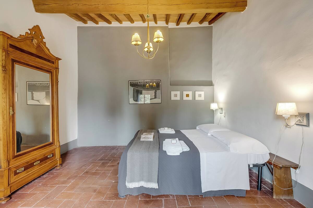 A bedroom with a bed, a wooden dresser, a mirror, a chandelier, and a wall-mounted light fixture.