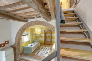 A room with a brick archway, wooden beams, a staircase, a radiator, a patterned rug, and a bed with a blue cover.