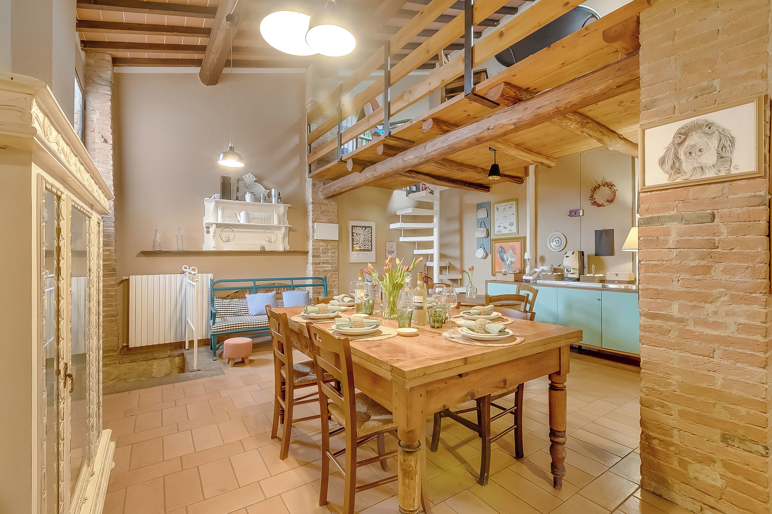 Een eetgedeelte met een houten tafel gedekt voor vier personen, een keuken op de achtergrond en een trap naar een hoger gelegen verdieping.