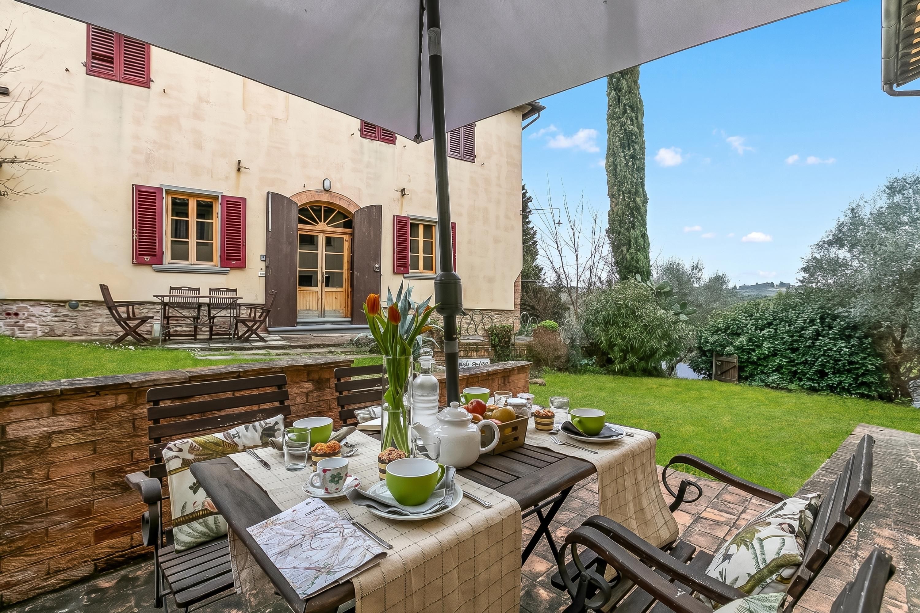 Un área de comedor al aire libre con una mesa preparada para dos, un banco y vista a un edificio con contraventanas rojas.