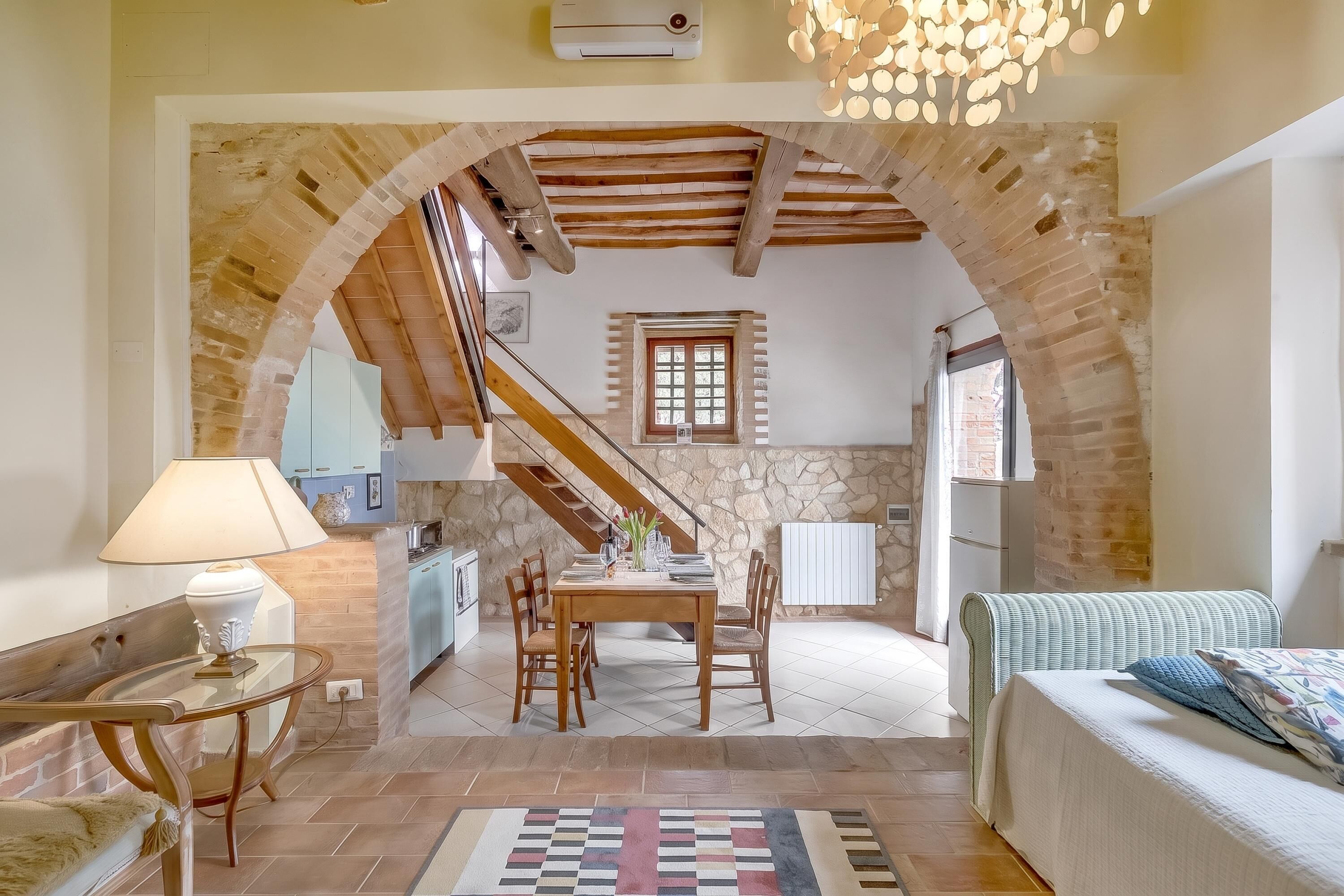Un interior acogedor con comedor, cocina y sala de estar, que presenta vigas de madera a la vista, paredes de piedra y suelo de baldosas.