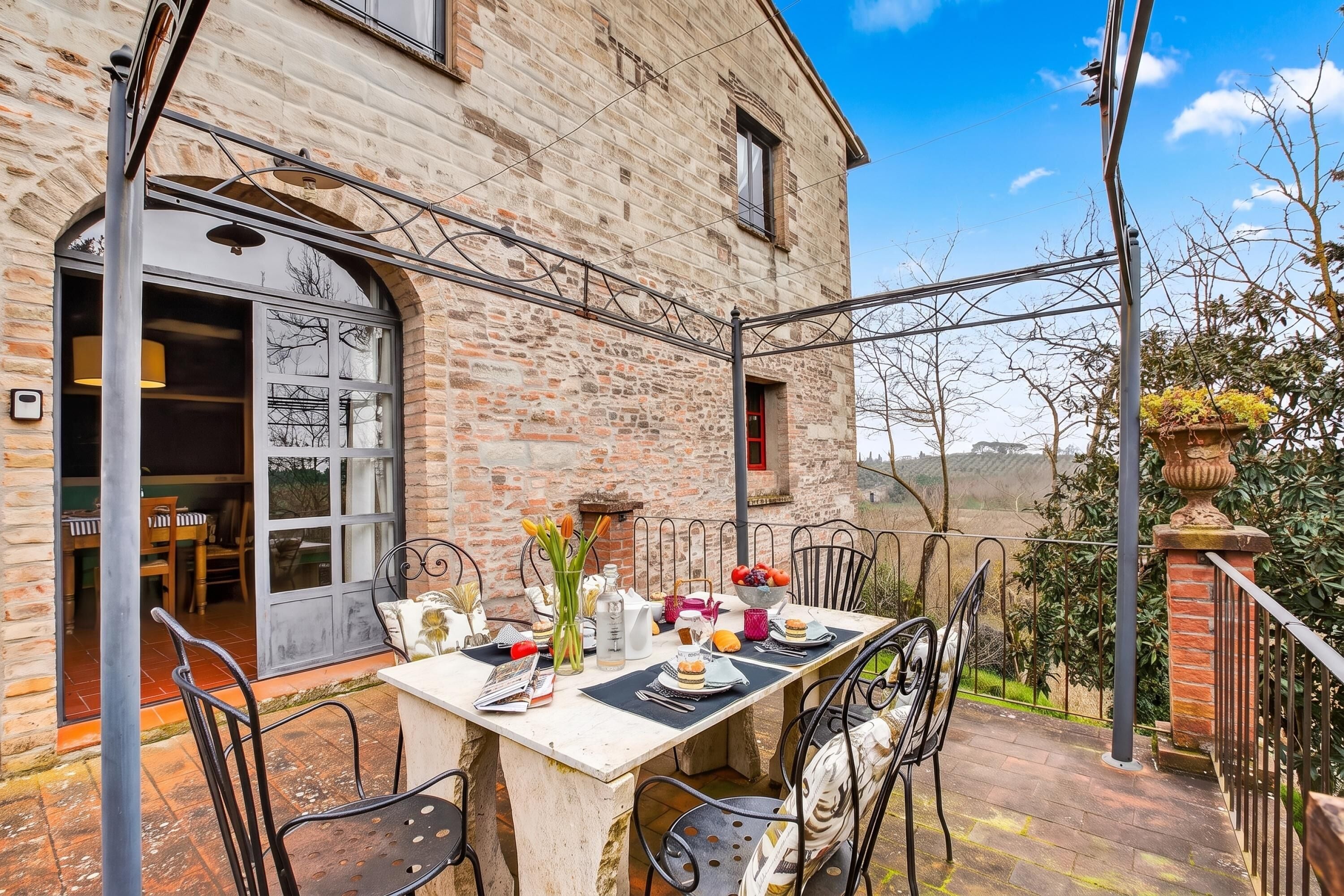 An outdoor dining area with a stone building, metal chairs, and a table set with plates, glasses, and flowers.