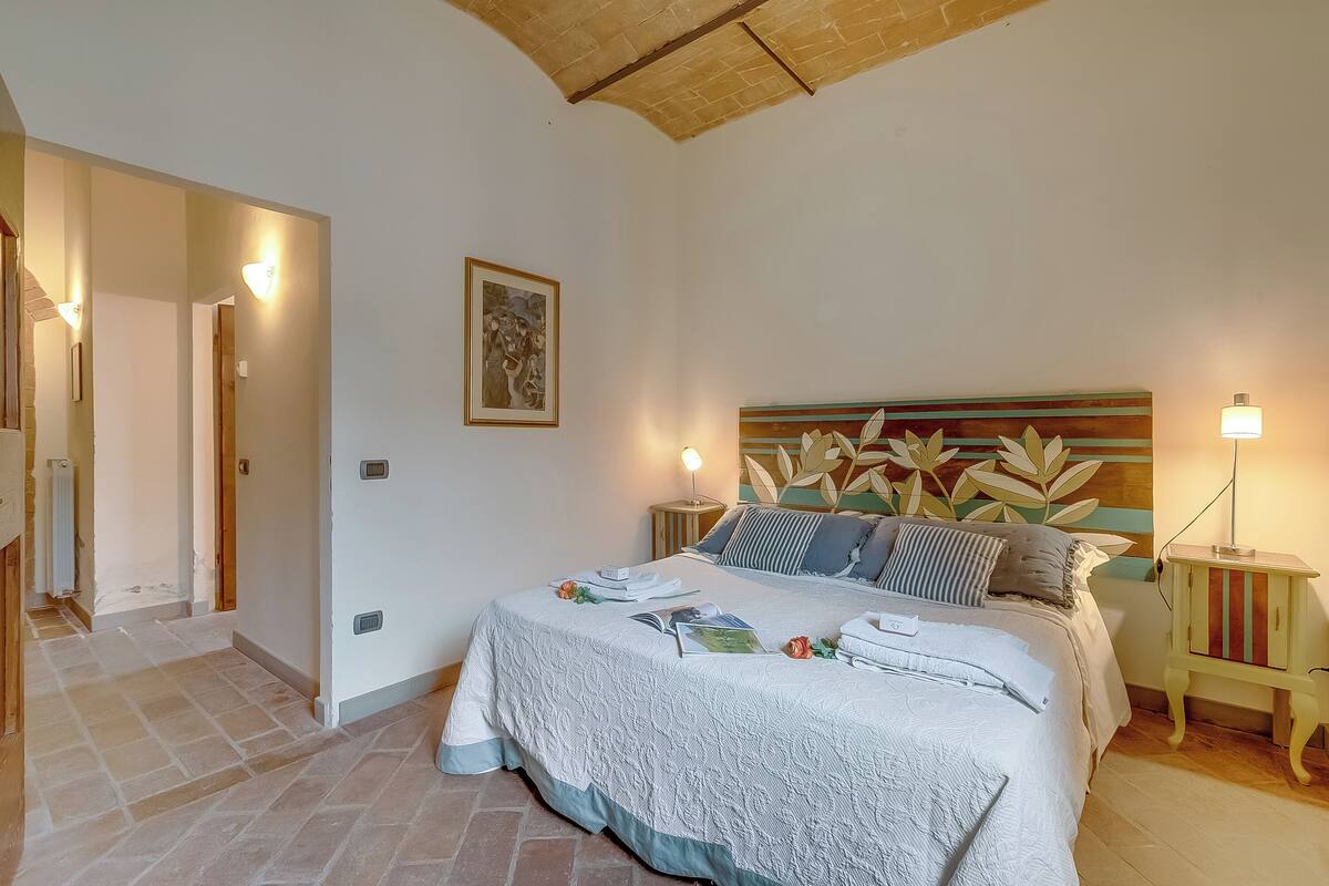 A bedroom with a large bed, bedside tables, a headboard with leaf patterns, a framed picture on the wall, and a small bedside lamp.