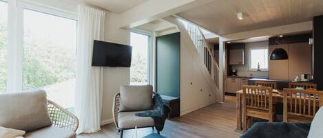 A modern living room with a sofa, armchair, and dining table. A staircase leads to a kitchen area.